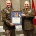 Two U.S. Army officers in service uniforms are posing together while holding a framed award certificate. The framed display contains a medal with a red ribbon and an official certificate reading 'The United States of America' and 'The Legion of Merit.' The ceremony appears to be a formal military awards presentation.