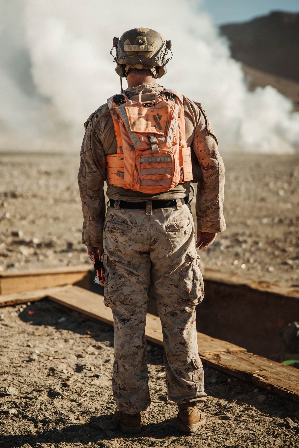 A military service member wearing USMC MARPAT desert camouflage uniform stands with their back to the camera, observing a large smoke cloud in an arid, desert-like environment. The individual is equipped with a tactical helmet with the name 'MCKINNE_' visible, and wears a distinctive bright orange safety/observer vest over their uniform. Wooden boards are visible on the ground nearby, suggesting a training range or demolition exercise area.
