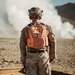 A military service member wearing USMC MARPAT desert camouflage uniform stands with their back to the camera, observing a large smoke cloud in an arid, desert-like environment. The individual is equipped with a tactical helmet with the name 'MCKINNE_' visible, and wears a distinctive bright orange safety/observer vest over their uniform. Wooden boards are visible on the ground nearby, suggesting a training range or demolition exercise area.