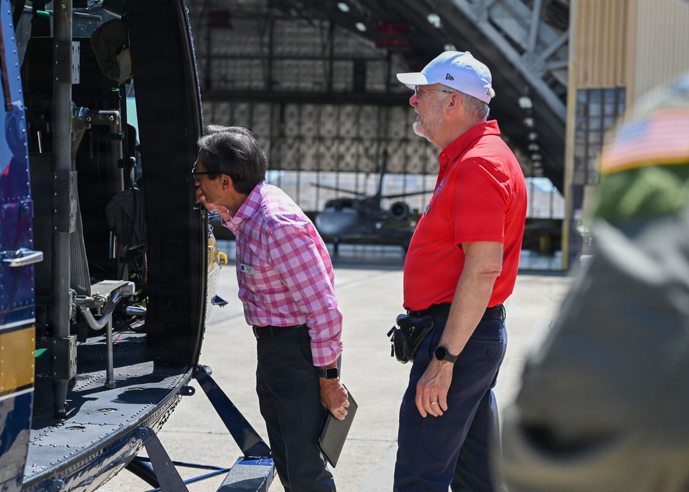 Two men are inspecting the interior of a helicopter on an airport tarmac, with a hangar visible in the background. One man in a pink checkered shirt is leaning in to look closely at the aircraft's interior, while the other in a red polo shirt and white cap observes. A jet aircraft is visible inside the hangar behind them.