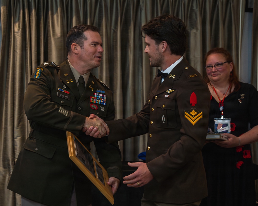 Two uniformed military personnel are shaking hands during what appears to be a formal award or recognition ceremony. The individual on the left, identified by a name tape reading 'JOSLYN', is presenting a framed item to the individual on the right, who wears insignia consistent with Canadian Armed Forces rank. A civilian woman wearing a lanyard badge stands in the background observing the exchange.