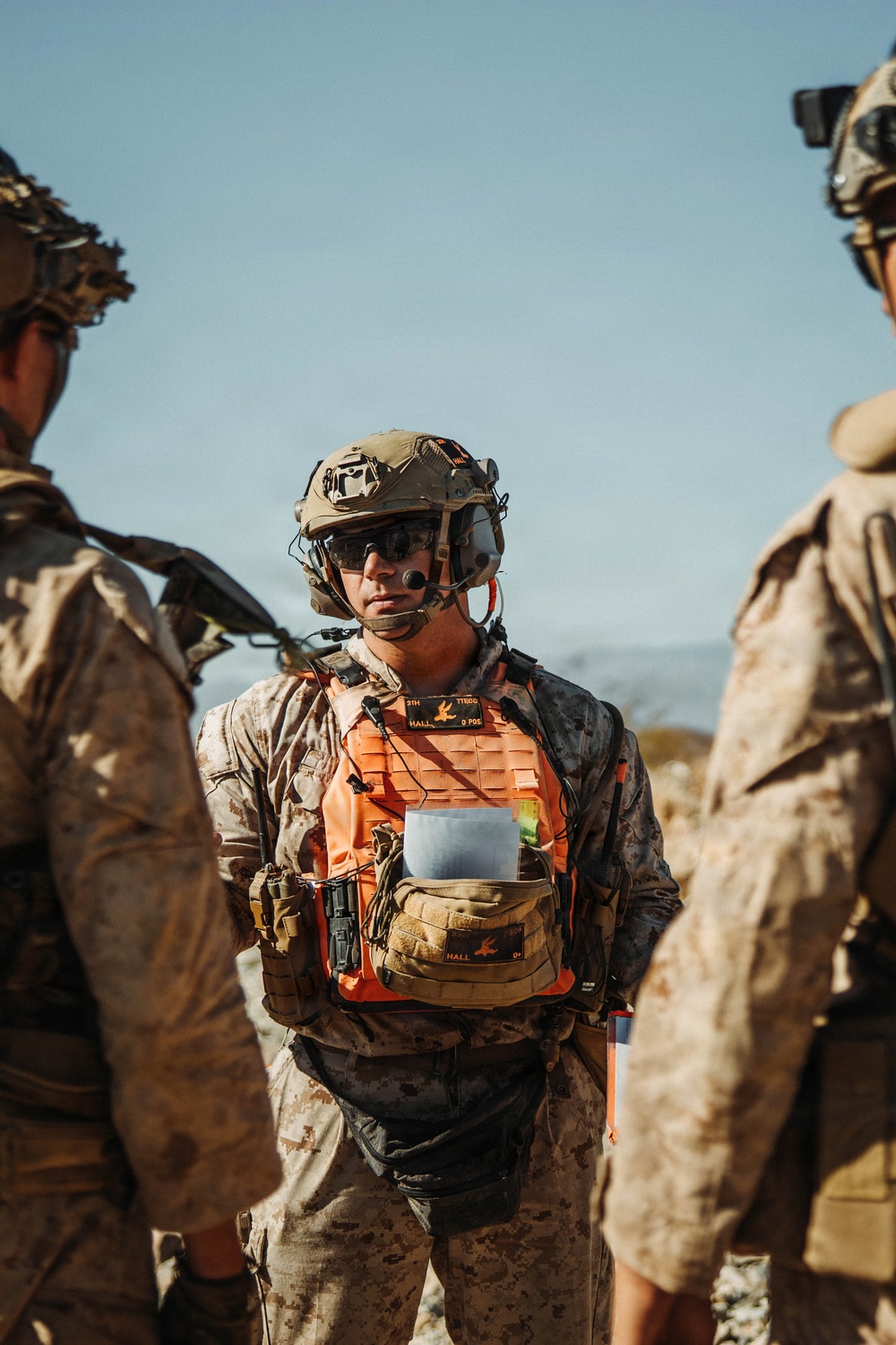 A military personnel wearing USMC desert MARPAT camouflage and a distinctive orange training/exercise vest is standing among other similarly dressed personnel in an arid outdoor environment. The central figure is equipped with a tactical helmet with communications gear, sunglasses, and a microphone, and is holding documents. The orange vest indicates a training exercise role, likely as a controller, observer, or safety officer.