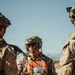 A military personnel wearing USMC desert MARPAT camouflage and a distinctive orange training/exercise vest is standing among other similarly dressed personnel in an arid outdoor environment. The central figure is equipped with a tactical helmet with communications gear, sunglasses, and a microphone, and is holding documents. The orange vest indicates a training exercise role, likely as a controller, observer, or safety officer.
