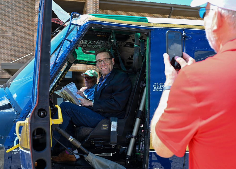 A smiling man in a dark blazer sits in the cockpit of a blue military helicopter, identified by visible U.S. Air Force markings and a serial number beginning with '69-6604'. A second person sits behind him holding papers, while a third person in a red shirt photographs them with a smartphone. The setting appears to be an outdoor public event or display near a brick building.