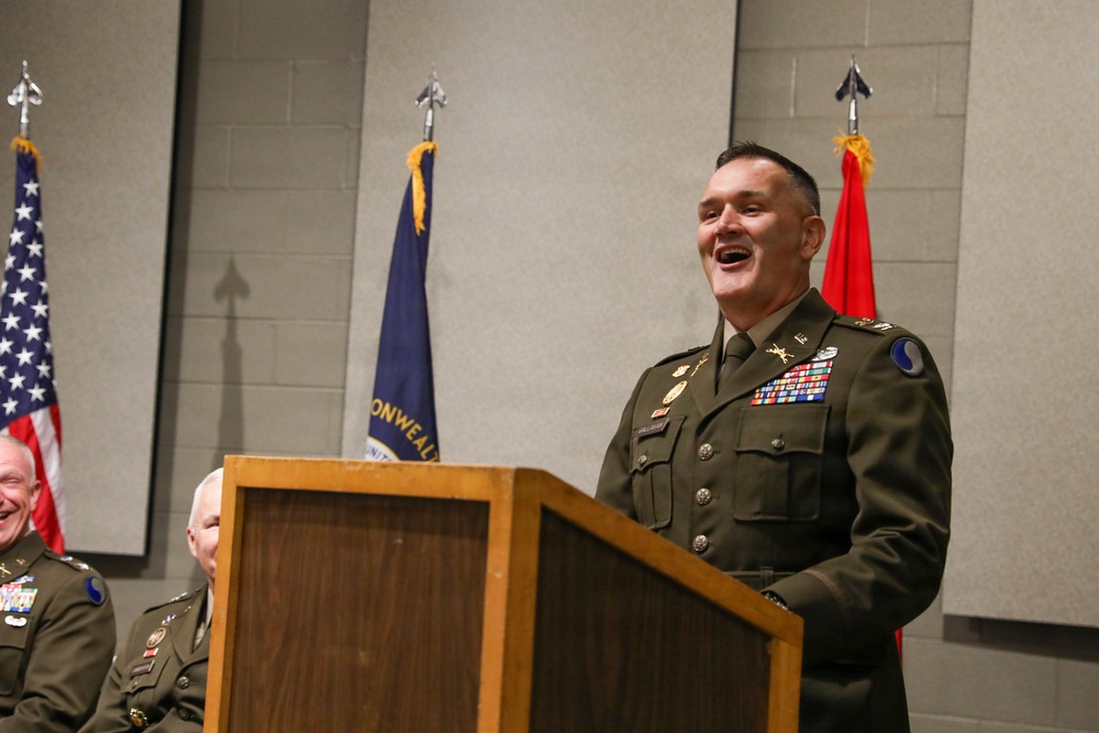 A U.S. Army officer in dress green uniform is speaking at a wooden podium, appearing to laugh or smile broadly during what appears to be a formal military ceremony. Two other uniformed officers are partially visible to the left. Three flags are displayed in the background, including the U.S. flag and what appears to be a Commonwealth state flag.