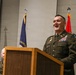 A U.S. Army officer in dress green uniform is speaking at a wooden podium, appearing to laugh or smile broadly during what appears to be a formal military ceremony. Two other uniformed officers are partially visible to the left. Three flags are displayed in the background, including the U.S. flag and what appears to be a Commonwealth state flag.