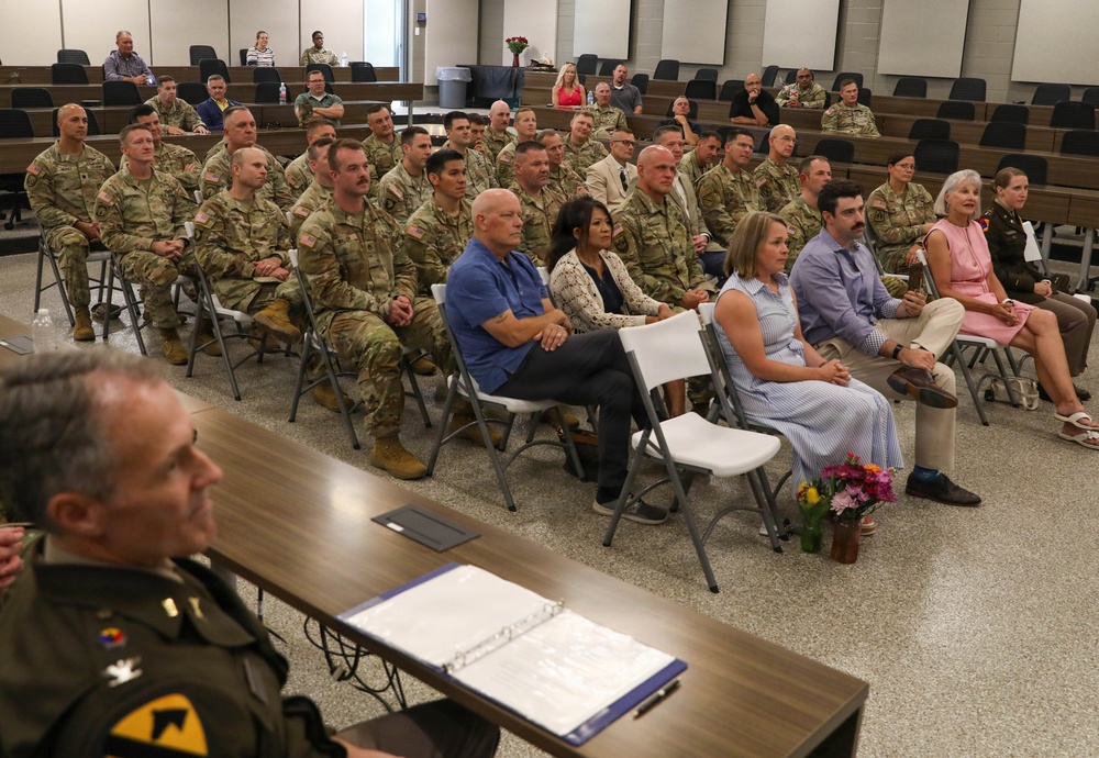 A military ceremony or briefing is taking place in an auditorium-style room, with a large audience consisting predominantly of uniformed soldiers in Army Combat Uniforms (ACUs) along with several civilians in casual attire. A senior officer in dress uniform is seated at a table in the foreground, facing the audience, with documents and a binder visible on the table. The setting suggests a formal military event such as a promotion ceremony, retirement, or unit briefing.