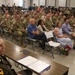 A military ceremony or briefing is taking place in an auditorium-style room, with a large audience consisting predominantly of uniformed soldiers in Army Combat Uniforms (ACUs) along with several civilians in casual attire. A senior officer in dress uniform is seated at a table in the foreground, facing the audience, with documents and a binder visible on the table. The setting suggests a formal military event such as a promotion ceremony, retirement, or unit briefing.