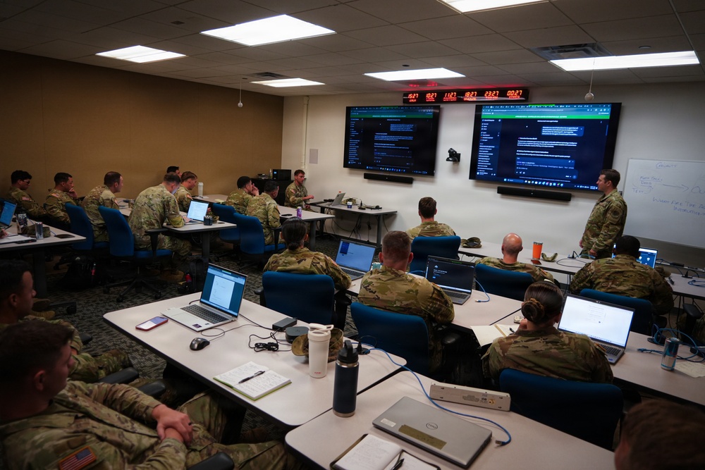 A military training or briefing session is taking place in a modern classroom environment. Numerous soldiers in camouflage uniforms are seated at tables with laptops while an instructor stands near two large display screens at the front of the room. A digital clock display showing multiple time zones is mounted above the screens, suggesting a coordinated or multi-theater operational context.