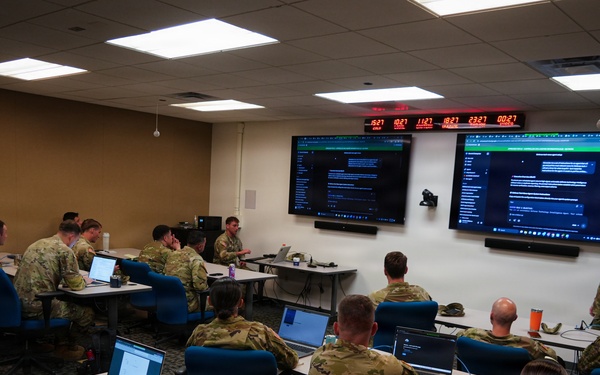 A military training or briefing session is taking place in a modern classroom environment. Numerous soldiers in camouflage uniforms are seated at tables with laptops while an instructor stands near two large display screens at the front of the room. A digital clock display showing multiple time zones is mounted above the screens, suggesting a coordinated or multi-theater operational context.