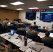 A military training or briefing session is taking place in a modern classroom environment. Numerous soldiers in camouflage uniforms are seated at tables with laptops while an instructor stands near two large display screens at the front of the room. A digital clock display showing multiple time zones is mounted above the screens, suggesting a coordinated or multi-theater operational context.