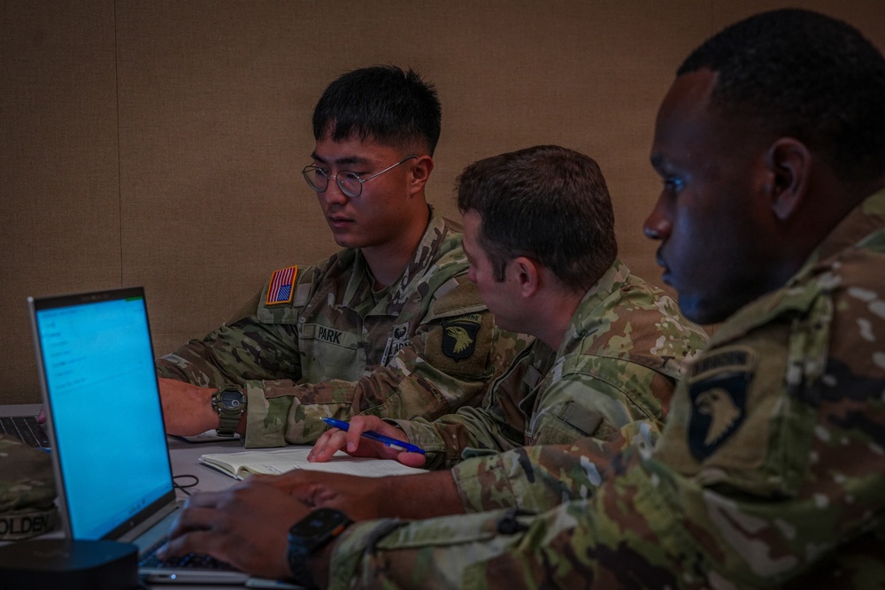 Three U.S. Army soldiers in Operational Camouflage Pattern (OCP) uniforms are seated at a table working collaboratively. They appear to be engaged in a planning or briefing session, with laptops open and one soldier taking notes. Visible name tapes and unit patches indicate affiliation with the 101st Airborne Division.