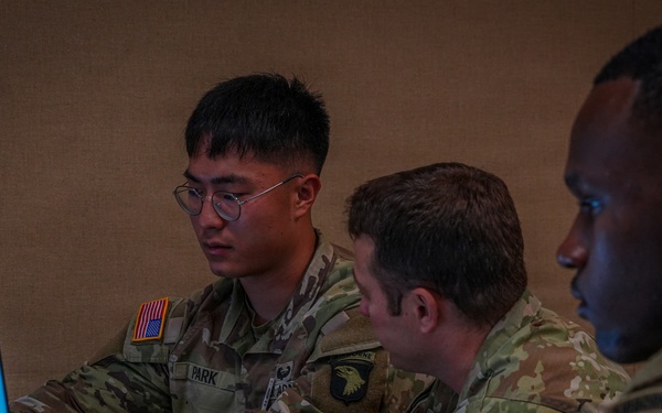Three U.S. Army soldiers in Operational Camouflage Pattern (OCP) uniforms are seated at a table working collaboratively. They appear to be engaged in a planning or briefing session, with laptops open and one soldier taking notes. Visible name tapes and unit patches indicate affiliation with the 101st Airborne Division.