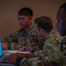 Three U.S. Army soldiers in Operational Camouflage Pattern (OCP) uniforms are seated at a table working collaboratively. They appear to be engaged in a planning or briefing session, with laptops open and one soldier taking notes. Visible name tapes and unit patches indicate affiliation with the 101st Airborne Division.