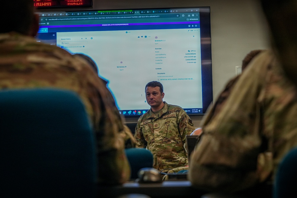 A U.S. Army soldier in OCP (Operational Camouflage Pattern) uniform appears to be conducting a briefing or training session in a classroom or operations room setting. A large monitor behind him displays what appears to be an AI platform interface, with visible text referencing 'Artificial Intelligence Strategy' and an agent labeled 'S6 Annex H'. Two other uniformed personnel are visible from behind in the foreground, seated as an audience.