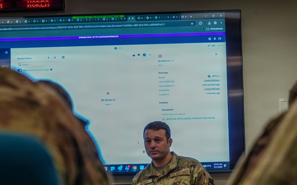 A U.S. Army soldier in OCP (Operational Camouflage Pattern) uniform appears to be conducting a briefing or training session in a classroom or operations room setting. A large monitor behind him displays what appears to be an AI platform interface, with visible text referencing 'Artificial Intelligence Strategy' and an agent labeled 'S6 Annex H'. Two other uniformed personnel are visible from behind in the foreground, seated as an audience.
