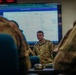 A U.S. Army soldier in OCP (Operational Camouflage Pattern) uniform appears to be conducting a briefing or training session in a classroom or operations room setting. A large monitor behind him displays what appears to be an AI platform interface, with visible text referencing 'Artificial Intelligence Strategy' and an agent labeled 'S6 Annex H'. Two other uniformed personnel are visible from behind in the foreground, seated as an audience.