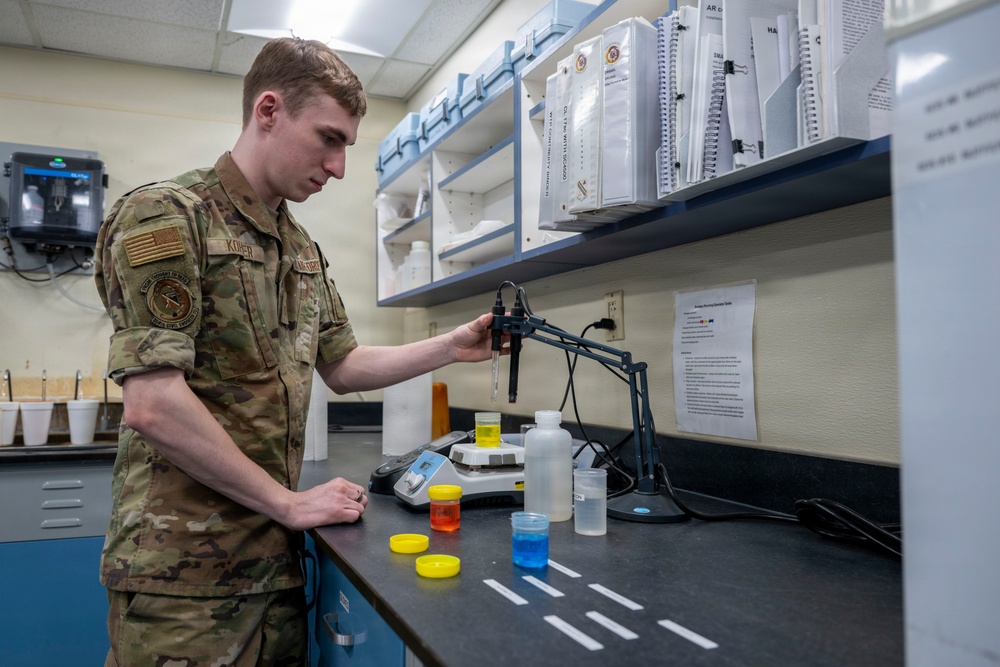 A U.S. military service member in MultiCam/OCP camouflage uniform is conducting laboratory work at a bench equipped with scientific instruments and colorful liquid samples. The individual appears to be operating a pipette or probe attached to a stand, likely performing water quality testing or chemical analysis. The setting is a well-organized military laboratory with reference binders, shelving, and various testing equipment.