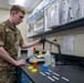 A U.S. military service member in MultiCam/OCP camouflage uniform is conducting laboratory work at a bench equipped with scientific instruments and colorful liquid samples. The individual appears to be operating a pipette or probe attached to a stand, likely performing water quality testing or chemical analysis. The setting is a well-organized military laboratory with reference binders, shelving, and various testing equipment.