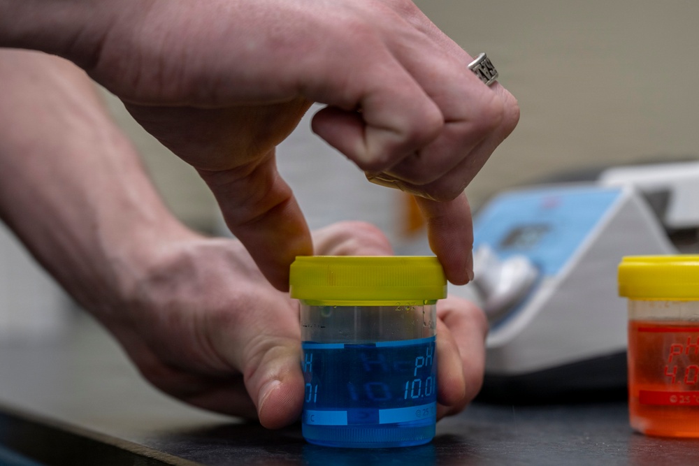 A person is handling a small laboratory container with a yellow lid containing blue liquid, likely a pH buffer solution with a visible pH value of 10.0 marked on the label. A second container with orange liquid and a yellow lid is visible in the foreground right. The setting appears to be a laboratory environment with analytical equipment visible in the background.