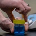 A person is handling a small laboratory container with a yellow lid containing blue liquid, likely a pH buffer solution with a visible pH value of 10.0 marked on the label. A second container with orange liquid and a yellow lid is visible in the foreground right. The setting appears to be a laboratory environment with analytical equipment visible in the background.