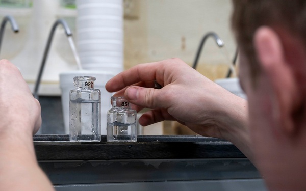 A person wearing military camouflage uniform is handling small labeled glass vials or bottles on a laboratory bench. Two clear glass bottles containing liquid are visible, with numeric labels on their caps. The setting appears to be a military or field laboratory environment with sinks visible in the background.