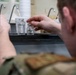 A person wearing military camouflage uniform is handling small labeled glass vials or bottles on a laboratory bench. Two clear glass bottles containing liquid are visible, with numeric labels on their caps. The setting appears to be a military or field laboratory environment with sinks visible in the background.