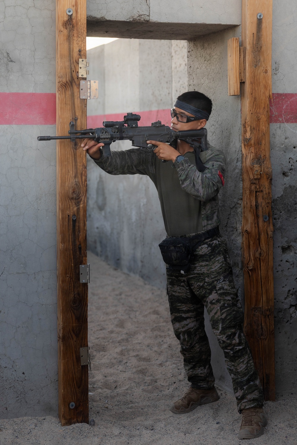 A soldier or trainee in camouflage uniform is performing a tactical door-clearing drill at what appears to be a military training facility. The individual is positioned in a doorway, aiming a rifle with an optical sight while using the door frame for cover. The setting features concrete walls with a red stripe marking, consistent with a mock urban combat training environment.
