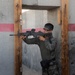 A soldier or trainee in camouflage uniform is performing a tactical door-clearing drill at what appears to be a military training facility. The individual is positioned in a doorway, aiming a rifle with an optical sight while using the door frame for cover. The setting features concrete walls with a red stripe marking, consistent with a mock urban combat training environment.