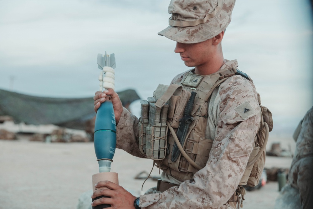 A U.S. Marine in MARPAT desert camouflage uniform and tactical vest is handling what appears to be a blue-painted training mortar round. The setting appears to be an arid, open training area. The blue color of the munition is consistent with inert/training ordnance used in military exercises.