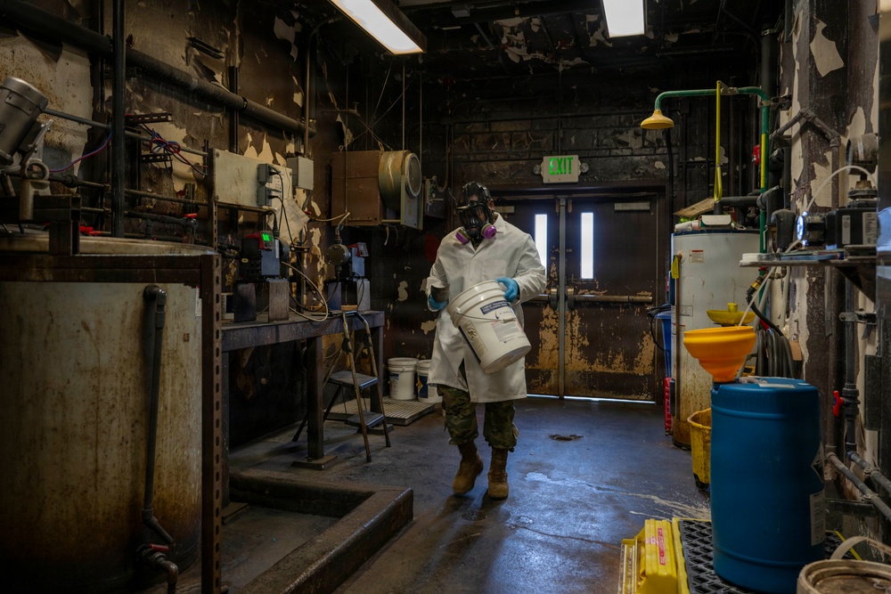 A worker in full personal protective equipment carries a white bucket through a heavily deteriorated industrial facility. The environment shows significant wear with peeling paint, corroded surfaces, and aged equipment. The setting appears to be a chemical handling or hazardous materials processing area.