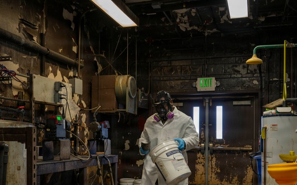 A worker in full personal protective equipment carries a white bucket through a heavily deteriorated industrial facility. The environment shows significant wear with peeling paint, corroded surfaces, and aged equipment. The setting appears to be a chemical handling or hazardous materials processing area.