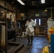 A worker in full personal protective equipment carries a white bucket through a heavily deteriorated industrial facility. The environment shows significant wear with peeling paint, corroded surfaces, and aged equipment. The setting appears to be a chemical handling or hazardous materials processing area.