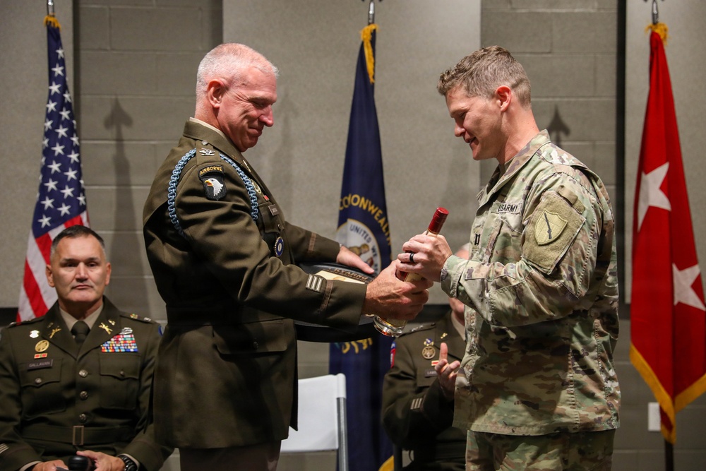 A military ceremony is taking place in which a senior officer in dress greens with an Airborne patch and multiple decorations is presenting an item to a soldier in Army Combat Uniform (ACU/OCP). A third officer in dress greens is seated in the background observing the ceremony. Flags including the U.S. flag, a state flag (Kentucky Commonwealth), and a general officer's flag (red with white star) are visible in the background.