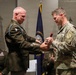 A military ceremony is taking place in which a senior officer in dress greens with an Airborne patch and multiple decorations is presenting an item to a soldier in Army Combat Uniform (ACU/OCP). A third officer in dress greens is seated in the background observing the ceremony. Flags including the U.S. flag, a state flag (Kentucky Commonwealth), and a general officer's flag (red with white star) are visible in the background.