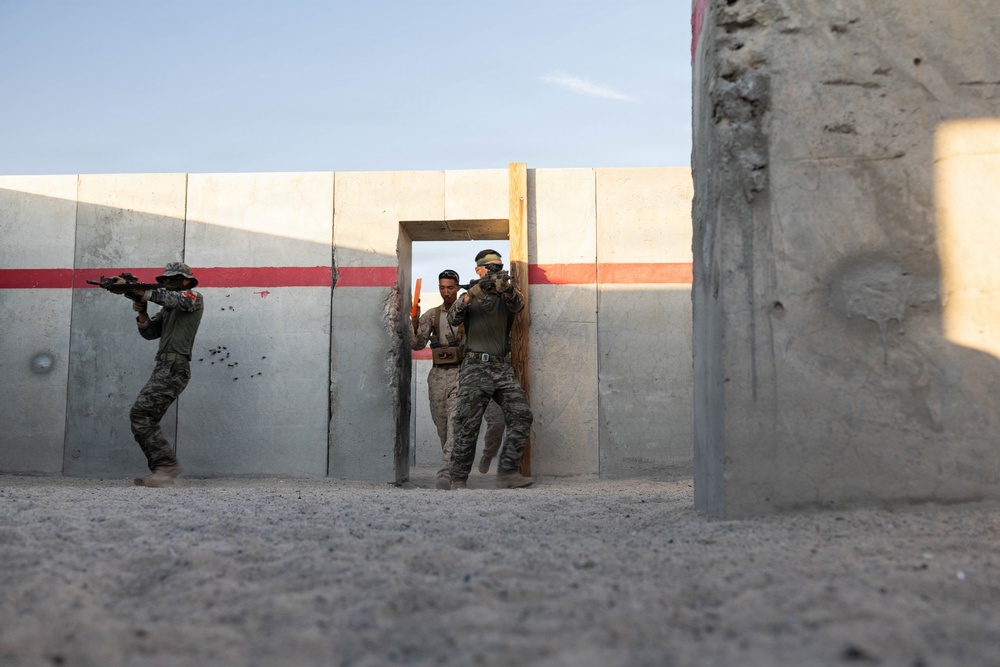 The image shows military or law enforcement personnel conducting a tactical training exercise at what appears to be a purpose-built urban warfare training facility. Three individuals in camouflage uniforms are performing a door-breach or room-clearing drill, with one positioned to the left of a concrete wall and two others moving through a doorway opening. The facility features concrete panel walls with a red stripe, simulating a built-up environment.