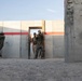 The image shows military or law enforcement personnel conducting a tactical training exercise at what appears to be a purpose-built urban warfare training facility. Three individuals in camouflage uniforms are performing a door-breach or room-clearing drill, with one positioned to the left of a concrete wall and two others moving through a doorway opening. The facility features concrete panel walls with a red stripe, simulating a built-up environment.