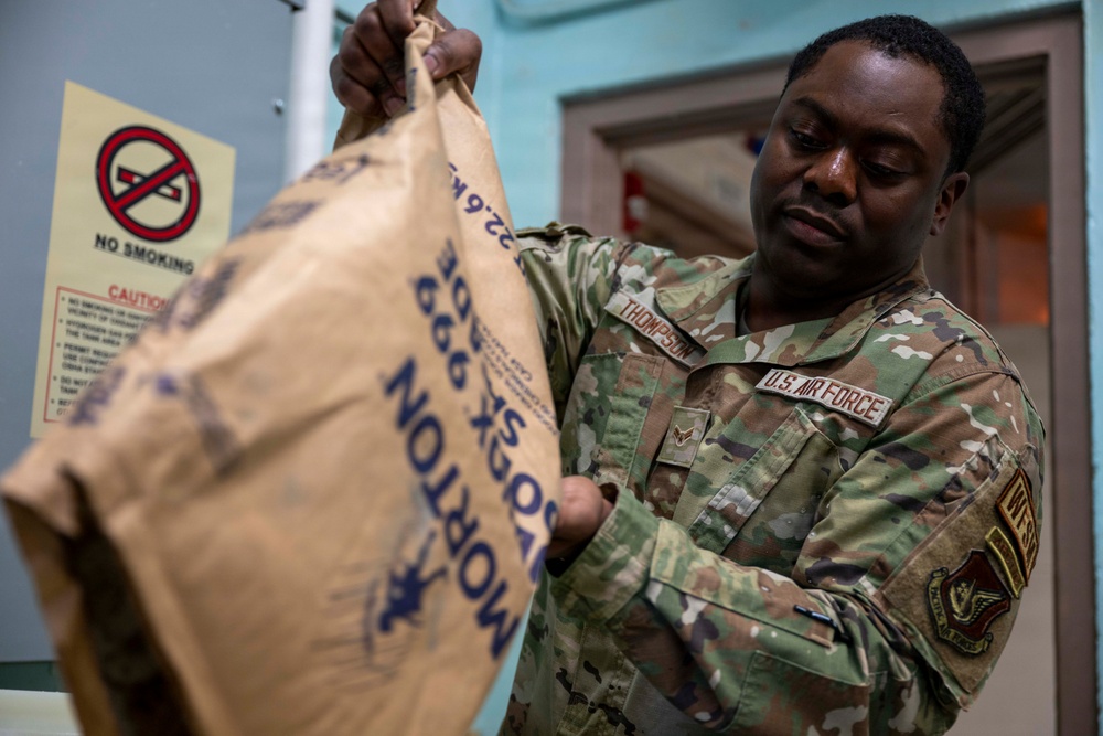 A U.S. Air Force service member wearing OCP (Operational Camouflage Pattern) uniform with the name tape 'Thompson' is handling a large Morton salt bag weighing 22.6 kg. The airman appears to be in an indoor facility, possibly performing a maintenance or logistics task involving the salt supply. A 'No Smoking' caution sign is visible on the wall in the background.
