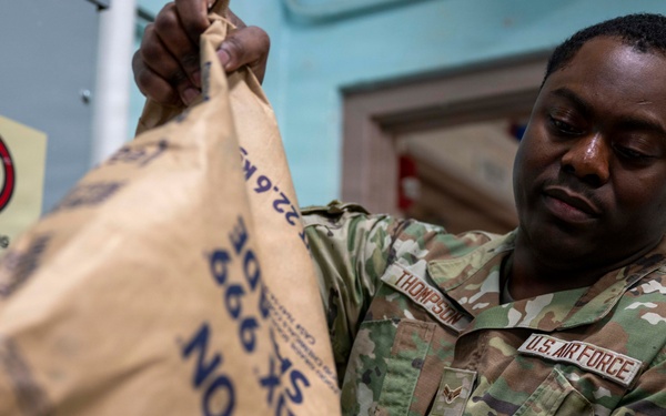 A U.S. Air Force service member wearing OCP (Operational Camouflage Pattern) uniform with the name tape 'Thompson' is handling a large Morton salt bag weighing 22.6 kg. The airman appears to be in an indoor facility, possibly performing a maintenance or logistics task involving the salt supply. A 'No Smoking' caution sign is visible on the wall in the background.