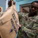 A U.S. Air Force service member wearing OCP (Operational Camouflage Pattern) uniform with the name tape 'Thompson' is handling a large Morton salt bag weighing 22.6 kg. The airman appears to be in an indoor facility, possibly performing a maintenance or logistics task involving the salt supply. A 'No Smoking' caution sign is visible on the wall in the background.