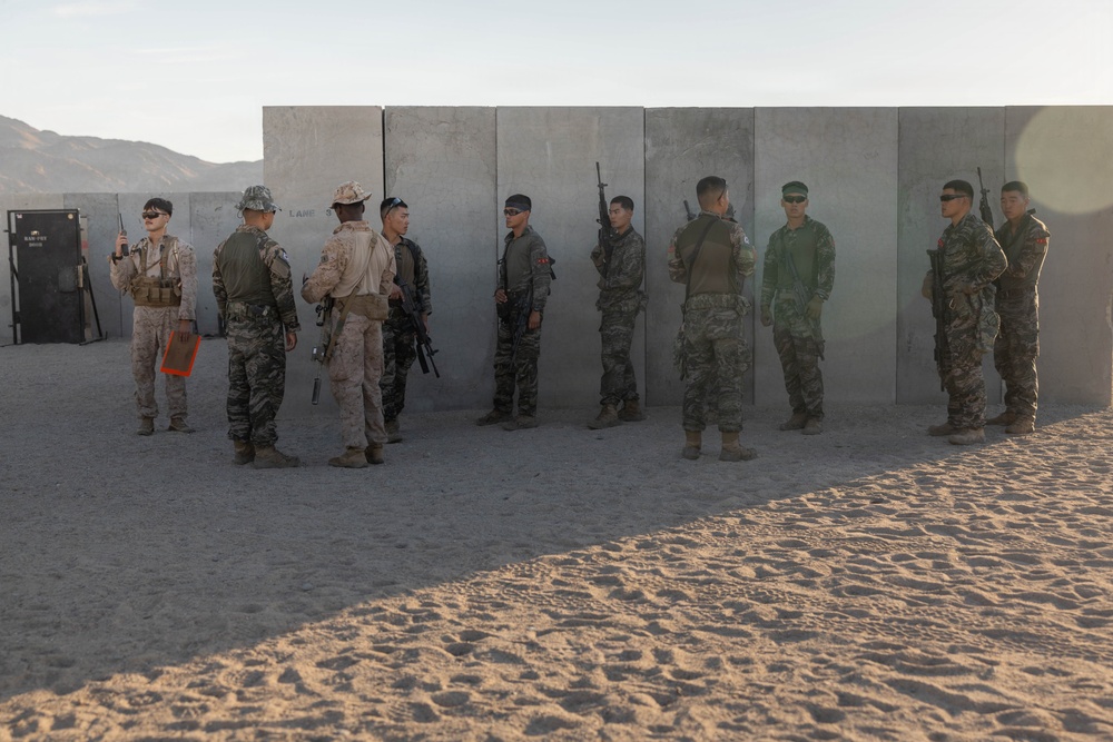 A group of approximately ten military personnel in various camouflage uniforms are gathered in front of a concrete wall structure in a desert training environment. The soldiers appear to be from multiple nations based on their differing uniform patterns and equipment. The setting suggests a joint military exercise or training range, likely in an arid region given the sandy terrain and mountain backdrop.