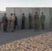 A group of approximately ten military personnel in various camouflage uniforms are gathered in front of a concrete wall structure in a desert training environment. The soldiers appear to be from multiple nations based on their differing uniform patterns and equipment. The setting suggests a joint military exercise or training range, likely in an arid region given the sandy terrain and mountain backdrop.