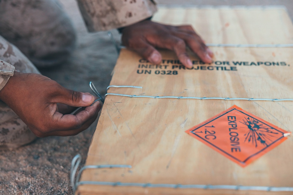 A person in military camouflage uniform is handling a wooden ammunition crate secured with wire. The crate is labeled with military ordnance markings indicating inert projectiles with UN classification 0328. An orange hazard diamond label marked 'EXPLOSIVE 1.2C' is affixed to the crate surface.