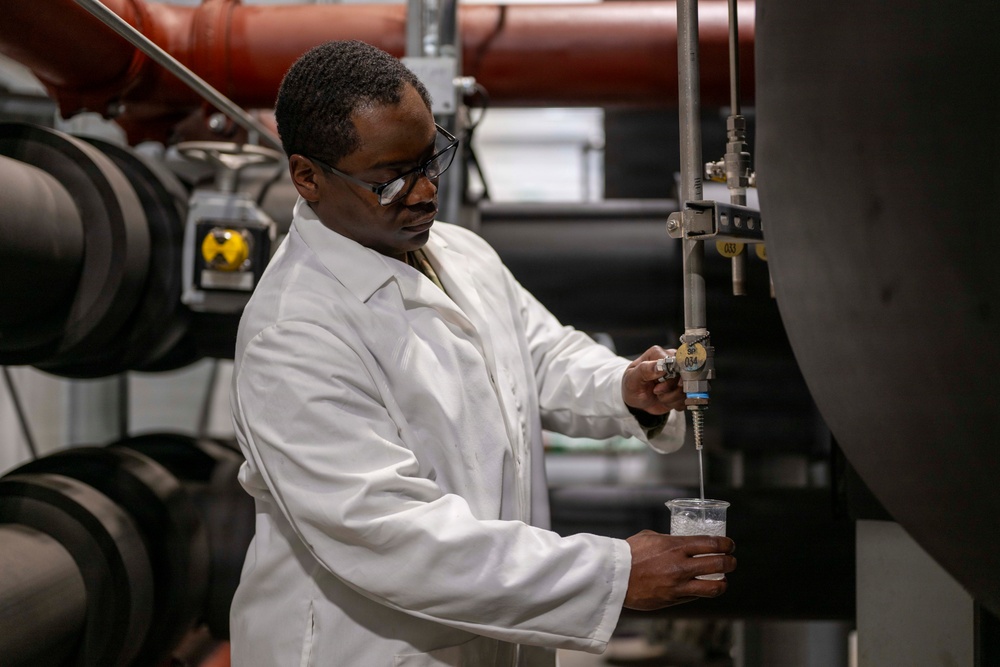 A laboratory technician or scientist wearing a white lab coat and safety glasses is collecting a sample from an industrial pipe or valve system. He is holding a clear plastic collection cup beneath a valve outlet, appearing to draw a fluid or gas sample. The setting appears to be an industrial facility or water treatment plant with large pipes and mechanical infrastructure visible in the background.