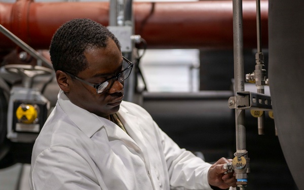 A laboratory technician or scientist wearing a white lab coat and safety glasses is collecting a sample from an industrial pipe or valve system. He is holding a clear plastic collection cup beneath a valve outlet, appearing to draw a fluid or gas sample. The setting appears to be an industrial facility or water treatment plant with large pipes and mechanical infrastructure visible in the background.