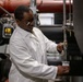 A laboratory technician or scientist wearing a white lab coat and safety glasses is collecting a sample from an industrial pipe or valve system. He is holding a clear plastic collection cup beneath a valve outlet, appearing to draw a fluid or gas sample. The setting appears to be an industrial facility or water treatment plant with large pipes and mechanical infrastructure visible in the background.