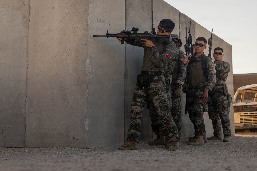 Four military personnel in camouflage uniforms are conducting a tactical training exercise at what appears to be a shooting range or urban warfare training facility. The lead soldier is aiming a rifle around the corner of a concrete wall while the others stack behind in a standard tactical formation. The setting includes concrete barrier walls marked 'LANE 3' and a derelict vehicle in the background, consistent with a military training environment.