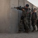 Four military personnel in camouflage uniforms are conducting a tactical training exercise at what appears to be a shooting range or urban warfare training facility. The lead soldier is aiming a rifle around the corner of a concrete wall while the others stack behind in a standard tactical formation. The setting includes concrete barrier walls marked 'LANE 3' and a derelict vehicle in the background, consistent with a military training environment.