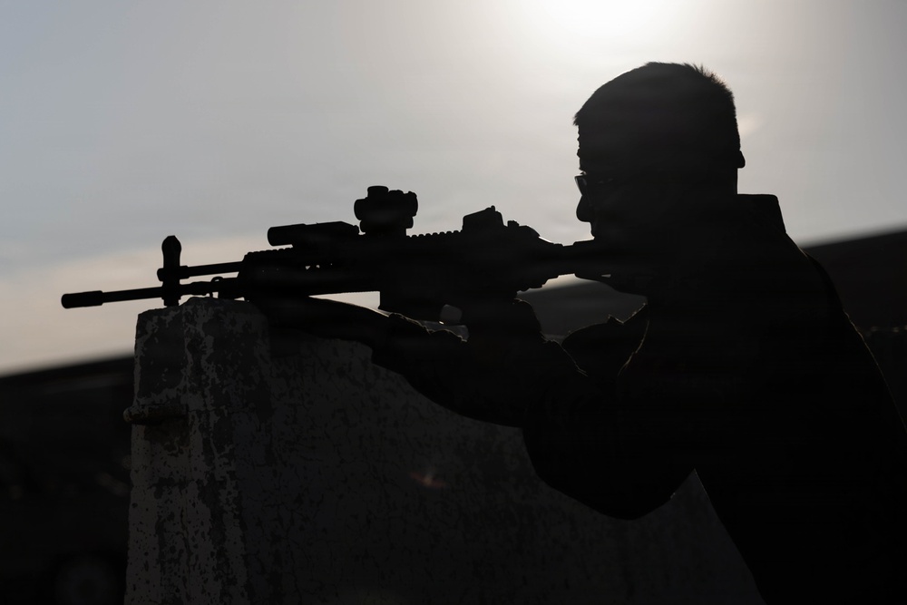 A silhouetted figure is positioned behind a concrete barrier, aiming a rifle equipped with optical sighting devices. The image is backlit by an overcast sky, rendering the subject almost entirely in shadow. The composition emphasizes the weapon's profile and the shooter's aiming stance.