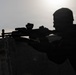 A silhouetted figure is positioned behind a concrete barrier, aiming a rifle equipped with optical sighting devices. The image is backlit by an overcast sky, rendering the subject almost entirely in shadow. The composition emphasizes the weapon's profile and the shooter's aiming stance.